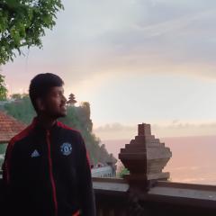 The image shows the author in black MUFC jacket with a temple and the sea in the background